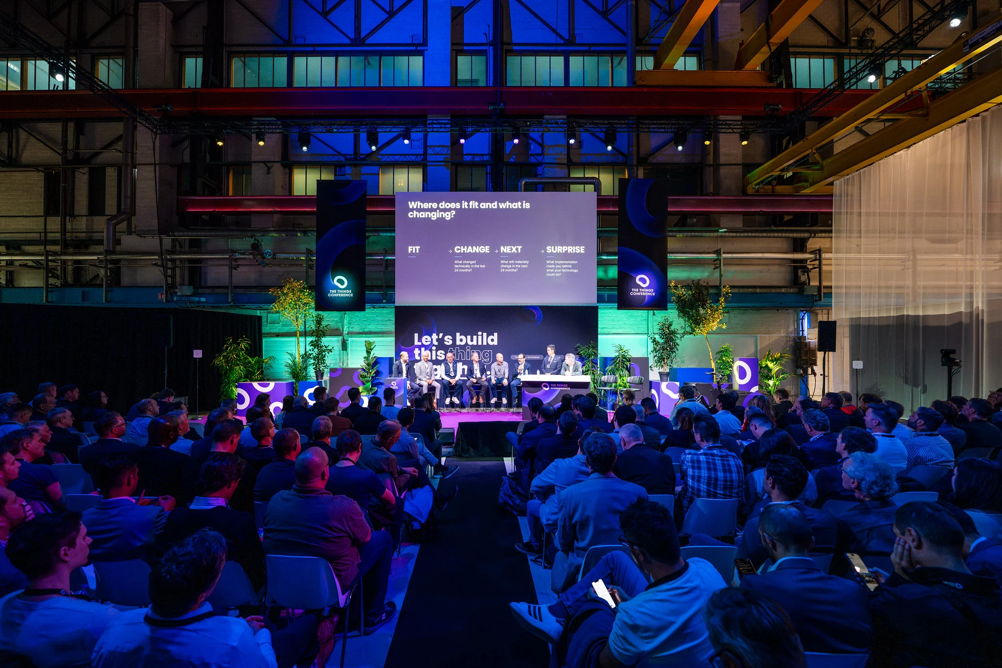 A full audience faces a panel of eight speakers on the Theater main stage, under a screen that reads "Where does it fit and what is changing?"